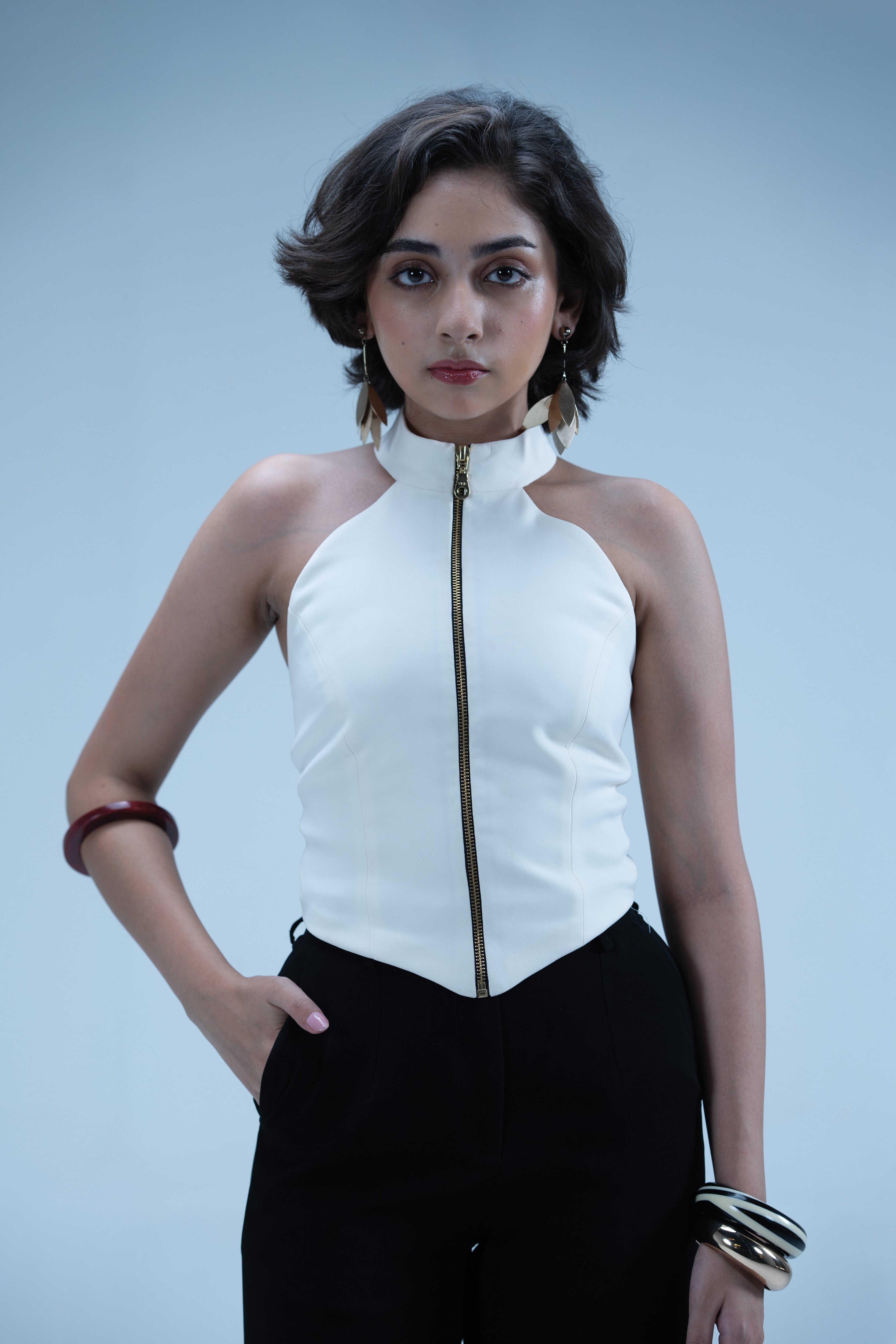 Woman wearing a white halter top with a zipper and black pants against a light blue background