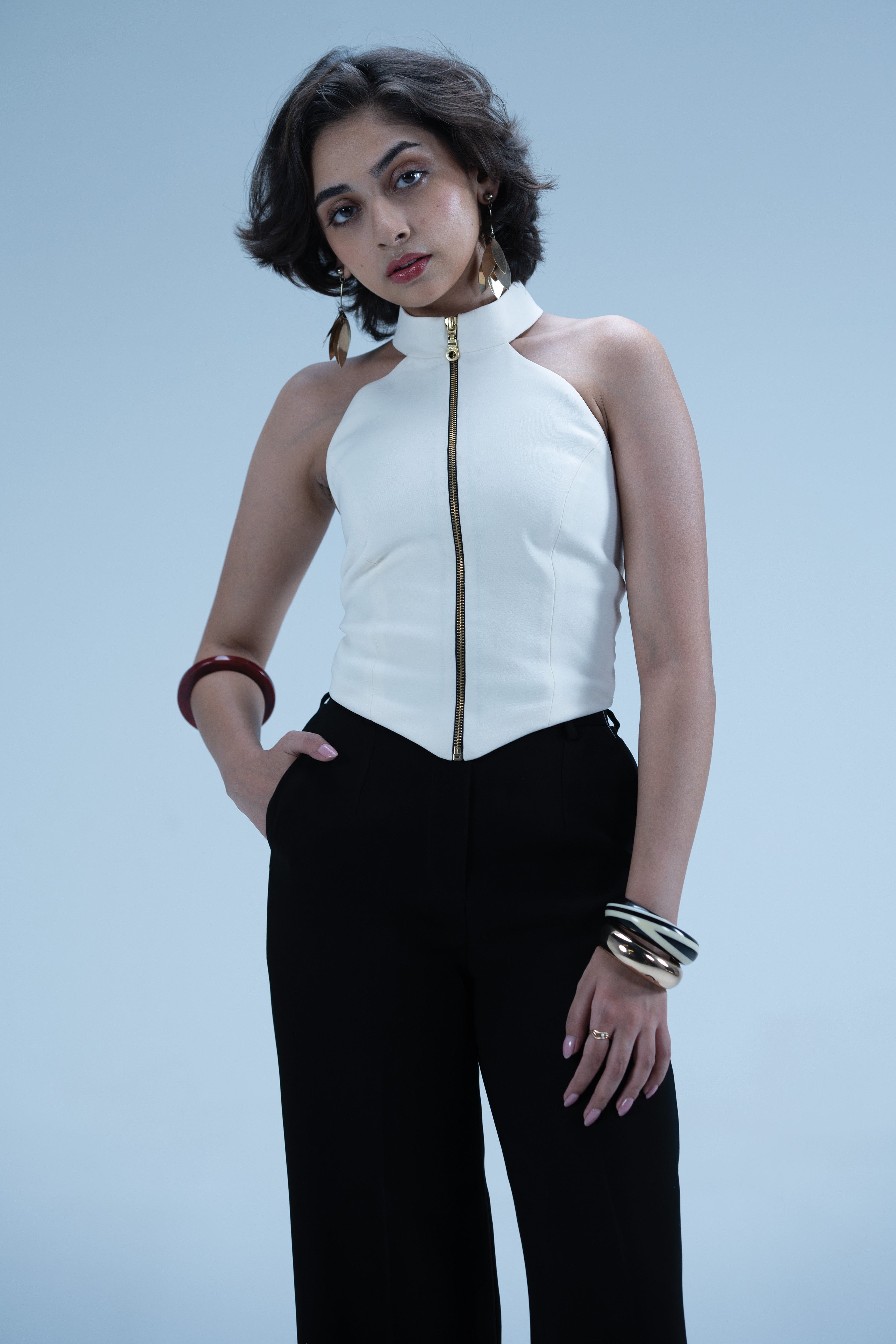 Woman wearing a white halter top with a gold zipper and black pants on a light blue background